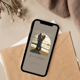Digitale Save-the-Date-Hochzeitskarte auf einem Smartphone, platziert auf beigem Stoff mit Trockenblumen, Umschlag und Papier. Auf dem Display eine elegante Save-the-Date-E-Card mit Hochzeitsfoto eines Paares am Strand bei Sonnenuntergang, Namen des Brautpaares und Hochzeitsdatum – stilvolle, moderne digitale Hochzeitseinladung.